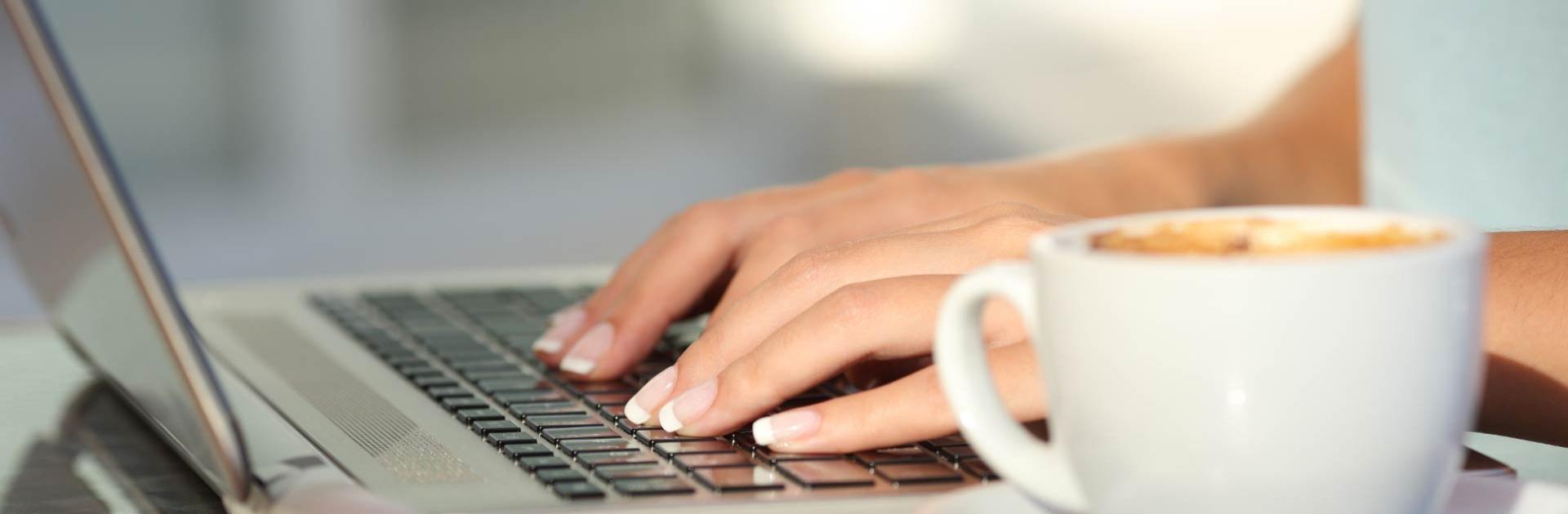 Mani che digitano su un laptop accanto a una tazza di caffè.