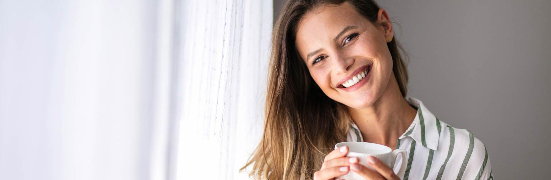 Donna sorridente con tazza, camicia a righe, vicino a una finestra.
